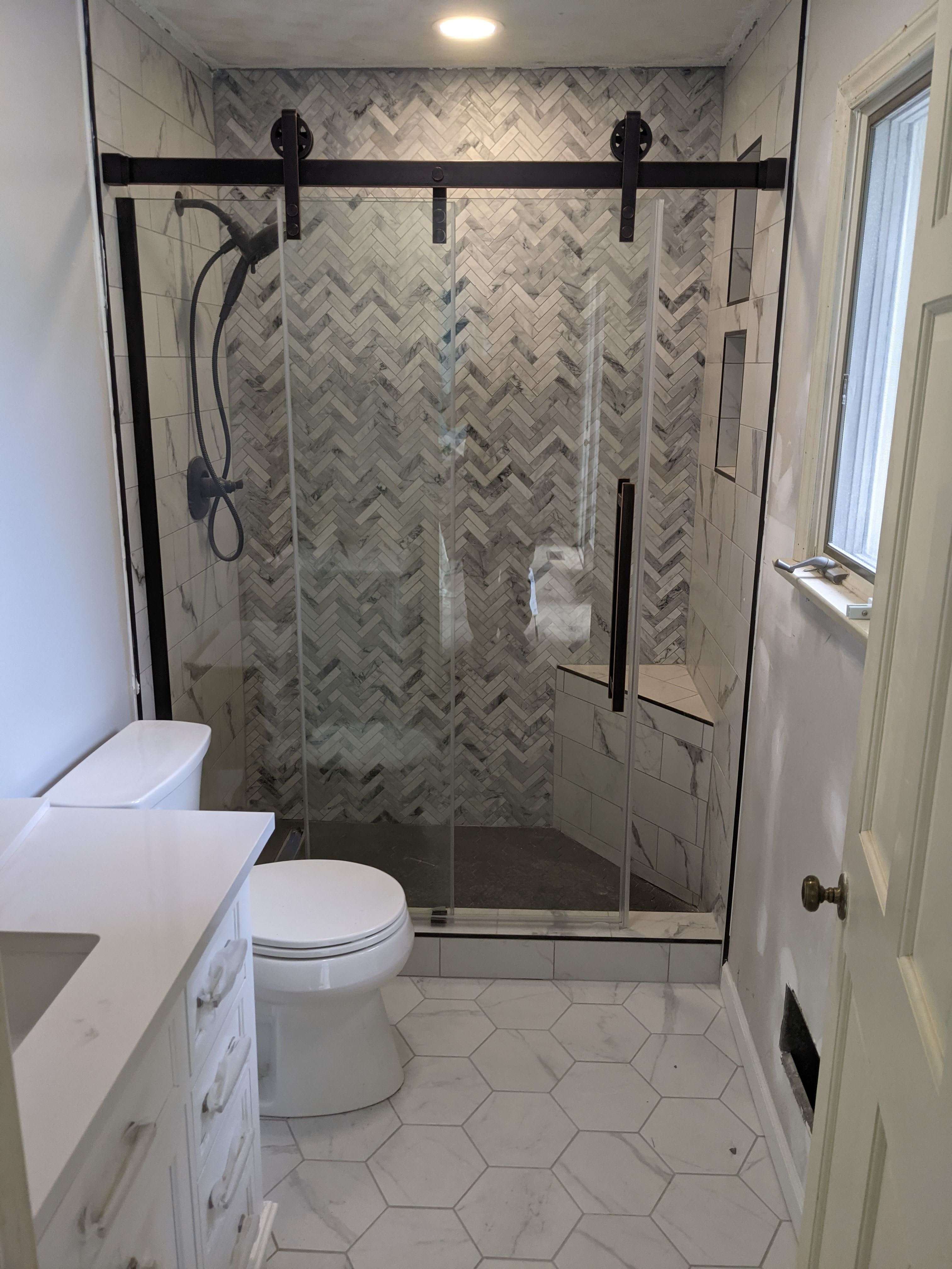 A modern renovated bathroom with a glass-enclosed walk-in shower, marble herringbone accent tiles, matte black barn-door hardware, and white hexagonal floor tiles.