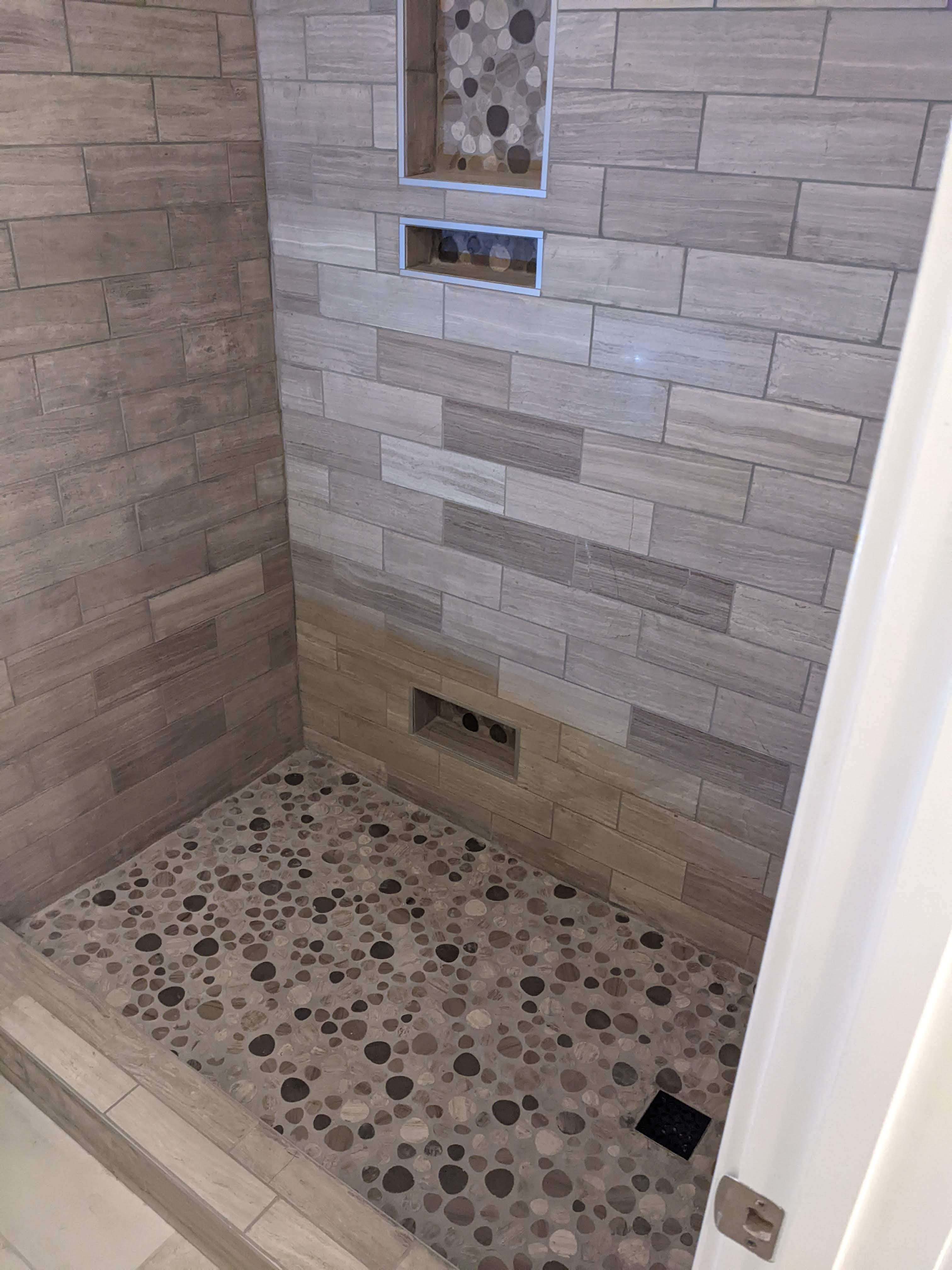 A custom walk-in shower featuring multi-toned gray and tan horizontal wall tiles, a pebble stone floor, and two built-in storage niches.