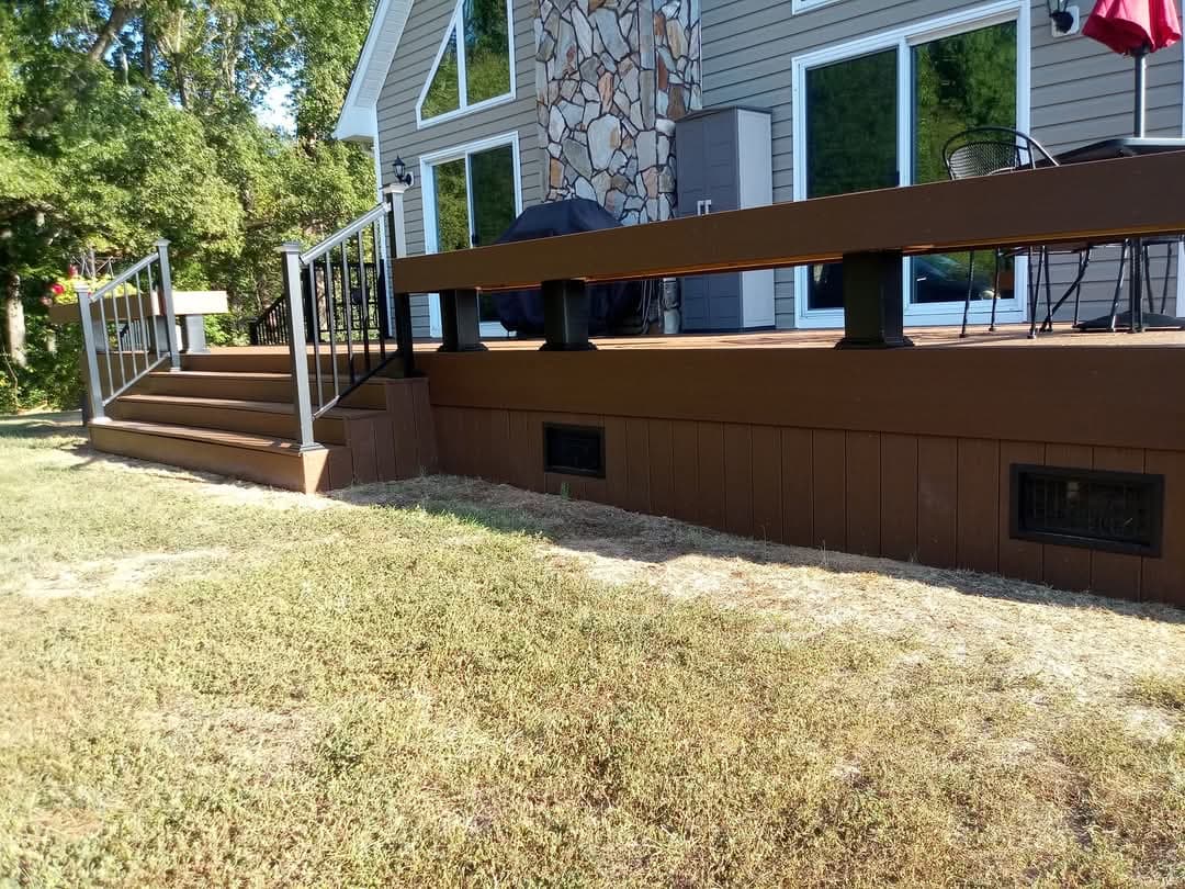 A completed brown composite deck with a dark railing system, integrated wide stairs, and a vertical skirting board with black vents.